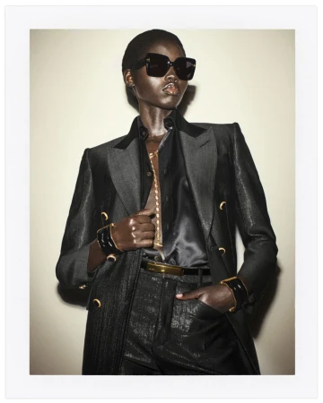 Fashion photo of Black female model in a dark suit with a black shirt, accessorized with gold jewelry and sunglasses, set against a plain background.