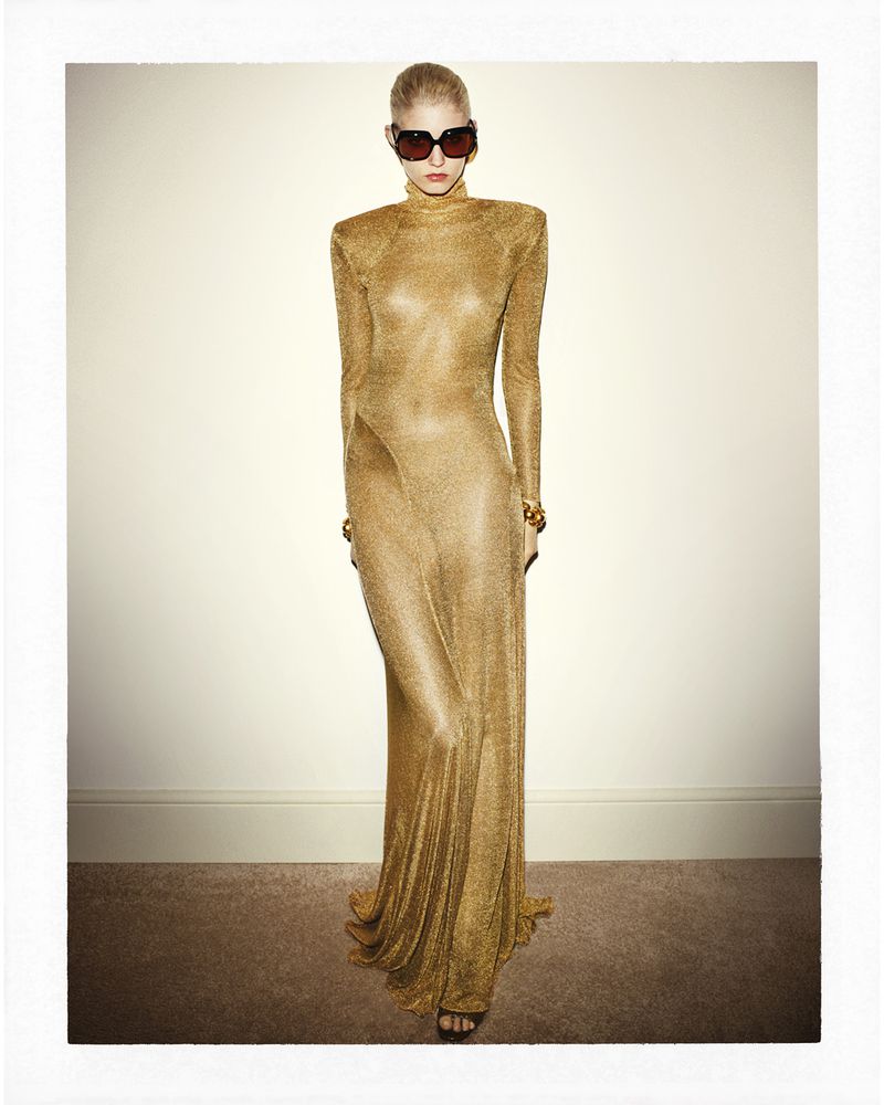 Elegant woman in a shimmering gold gown poses in front of a light beige wall wearing sunglasses and golden bracelets in a studio setting.