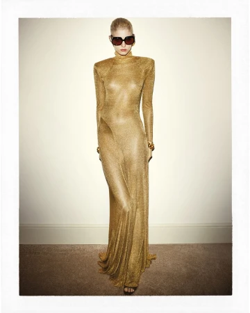 Elegant woman in a shimmering gold gown poses in front of a light beige wall wearing sunglasses and golden bracelets in a studio setting.