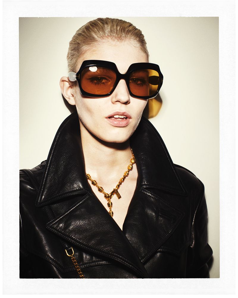Blonde woman with a black leather jacket and orange sunglasses, wearing a gold necklace, poses indoors against an off-white background.