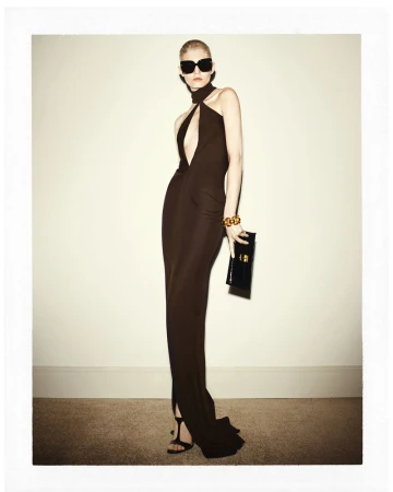 Model wearing a dark cutout evening dress, sunglasses, and a gold cuff against a plain studio background.