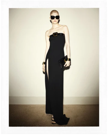 Model wearing oversized black sunglasses and a strapless black gown with a high slit, holding a clutch against a plain wall.