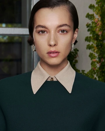 Portrait of a young woman with short dark hair, wearing a dark green top with a light beige collar, posing in front of a window.