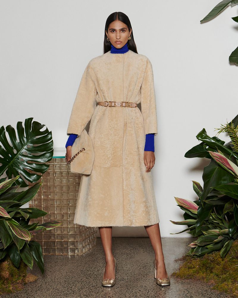 A woman wearing a beige coat, blue turtleneck, and gold heels stands in front of tropical plants holding a matching beige purse.