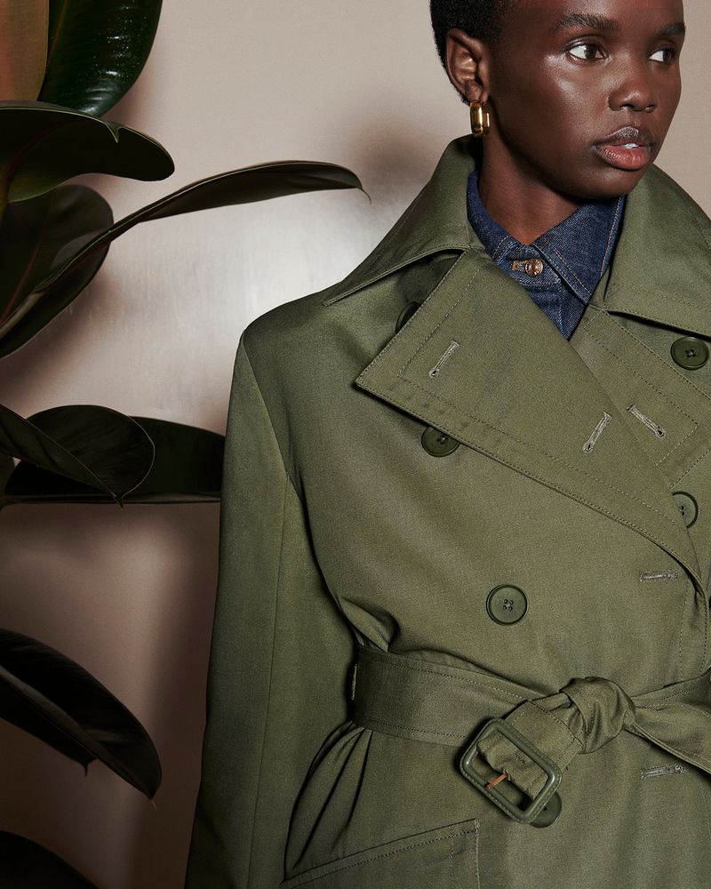A woman models a double-breasted trench coat in olive green, featuring a tie waist and large lapel in a studio setting with plant.
