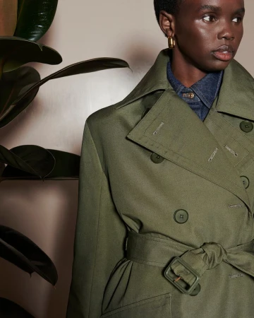 A woman models a double-breasted trench coat in olive green, featuring a tie waist and large lapel in a studio setting with plant.