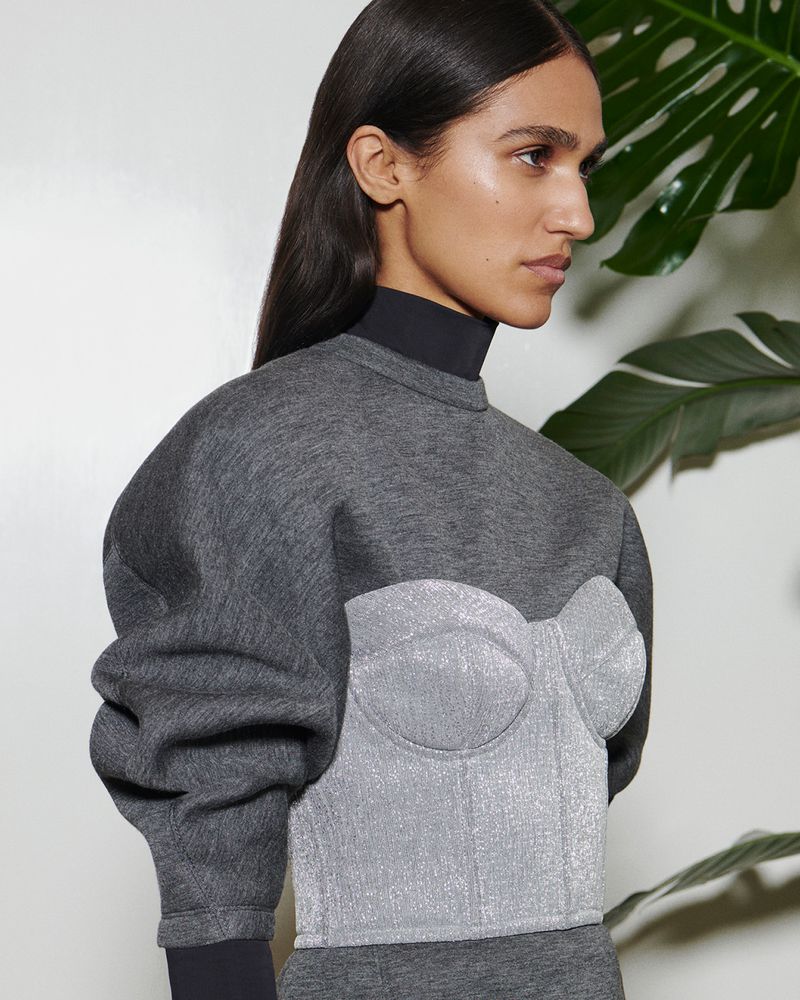 A stylish adult female poses indoors in a gray sweater with a silver corset over it, complemented by a monstera leaf in the background.