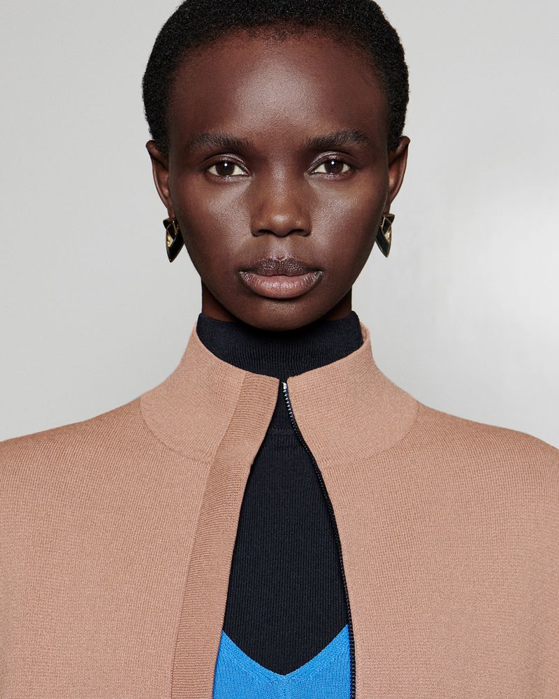 Close-up portrait of a dark-skinned Black woman wearing a tan cardigan over a black turtleneck and a blue shirt. She has black earrings.