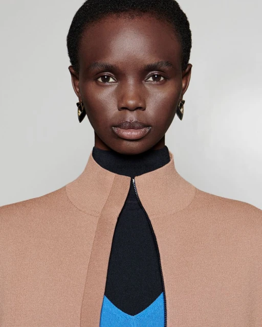 Close-up portrait of a dark-skinned Black woman wearing a tan cardigan over a black turtleneck and a blue shirt. She has black earrings.