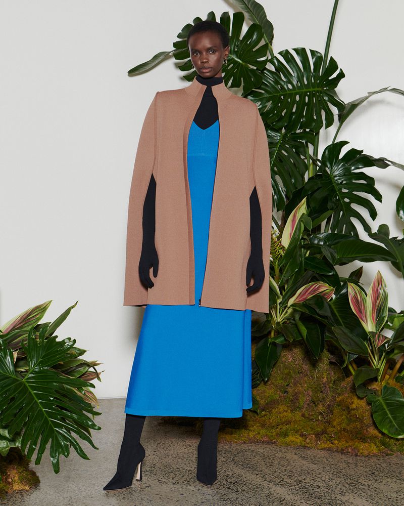 A black female fashion model poses in a studio wearing a beige cape, a bright blue dress, and black boots amid a leafy green backdrop.