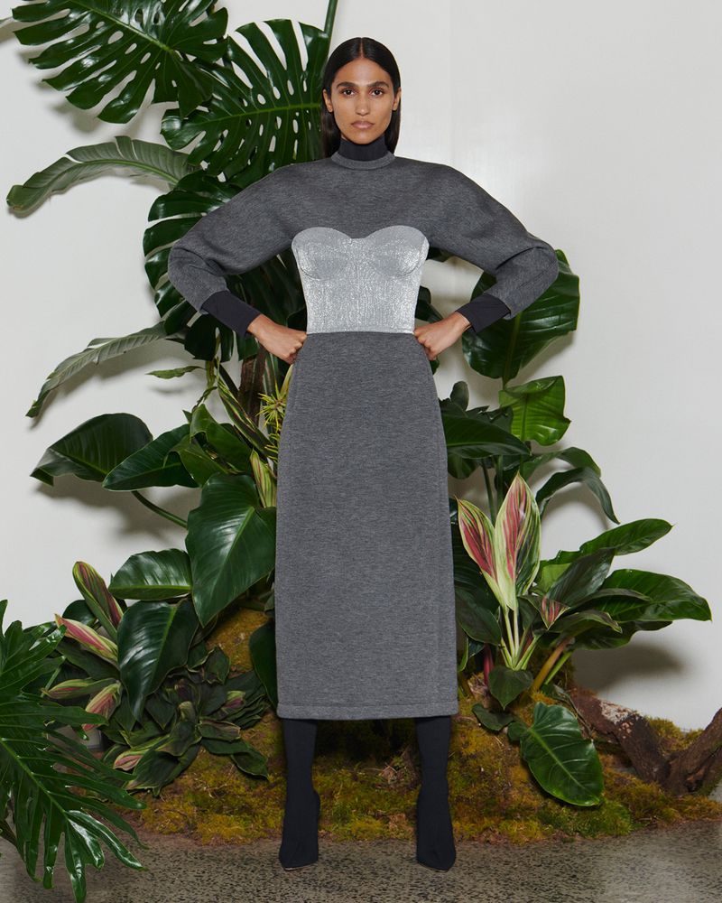 A woman in a gray knit dress with a sparkly silver bustier poses against a backdrop of lush green tropical foliage in a studio setting.