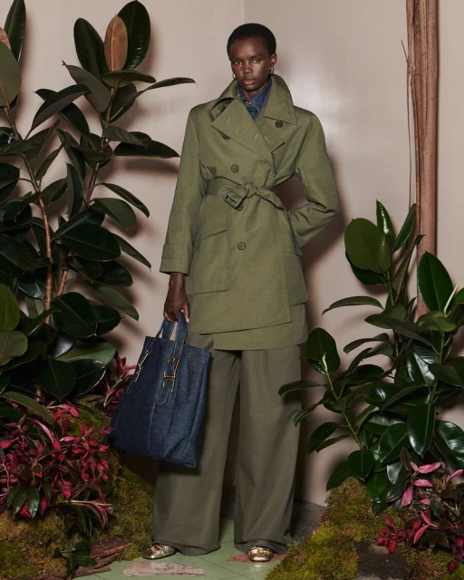 A woman in a green trench coat and wide-legged trousers holds a tote bag, surrounded by lush foliage in a studio setting.