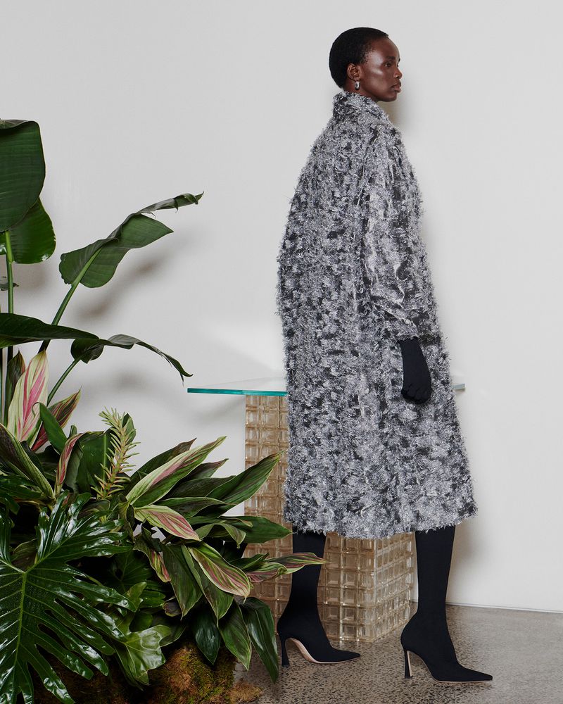 Full-length shot of a black woman in a patterned grey coat, black gloves, black socks and heeled shoes posing next to green leafy plants.