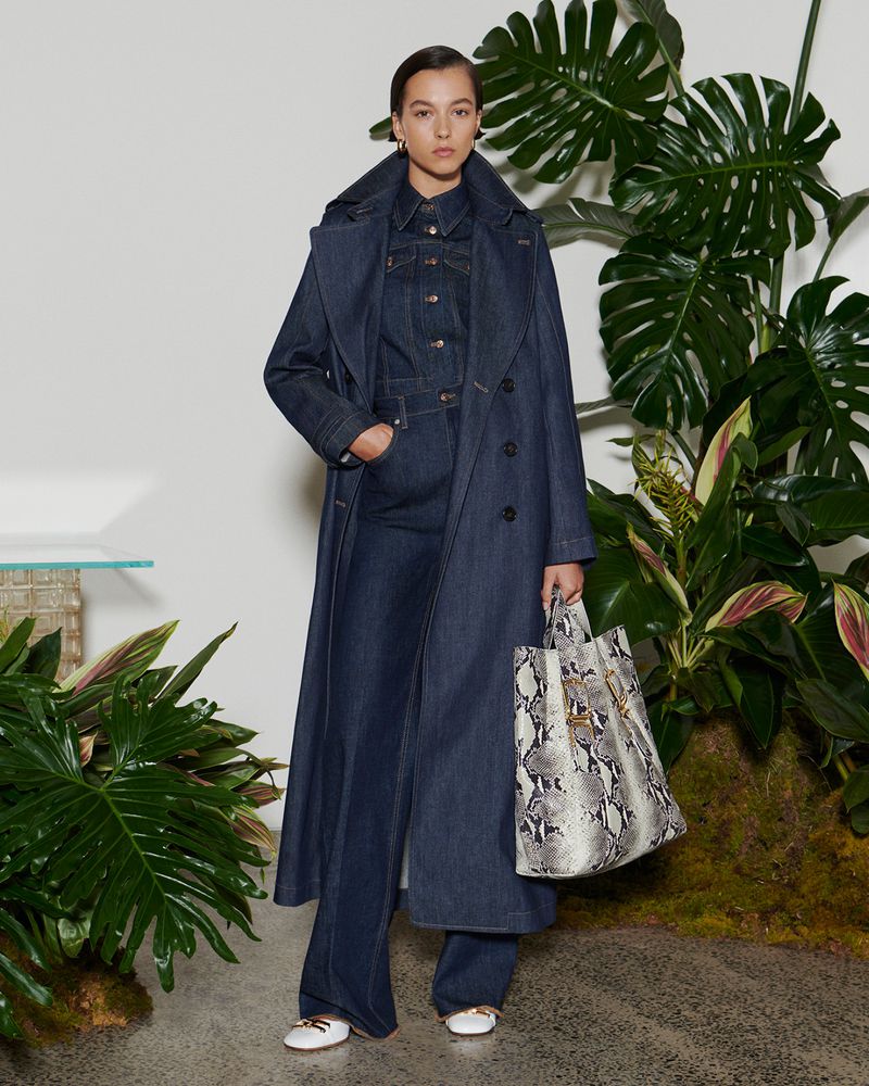 A woman in a blue denim trench coat and flared jeans stands in front of a backdrop of plants holding a snakeskin print tote bag.