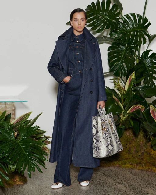 A woman in a blue denim trench coat and flared jeans stands in front of a backdrop of plants holding a snakeskin print tote bag.