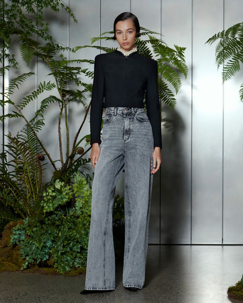 A young adult woman poses in a black jacket and grey wide-leg jeans against a backdrop of ferns and a reflective panel in a studio setting.