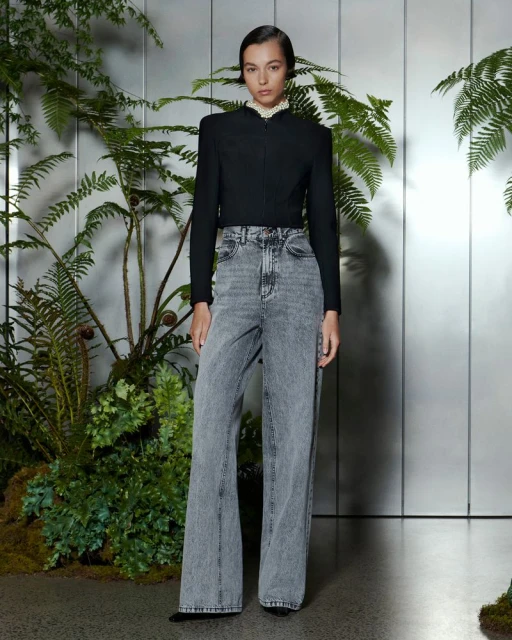 A young adult woman poses in a black jacket and grey wide-leg jeans against a backdrop of ferns and a reflective panel in a studio setting.