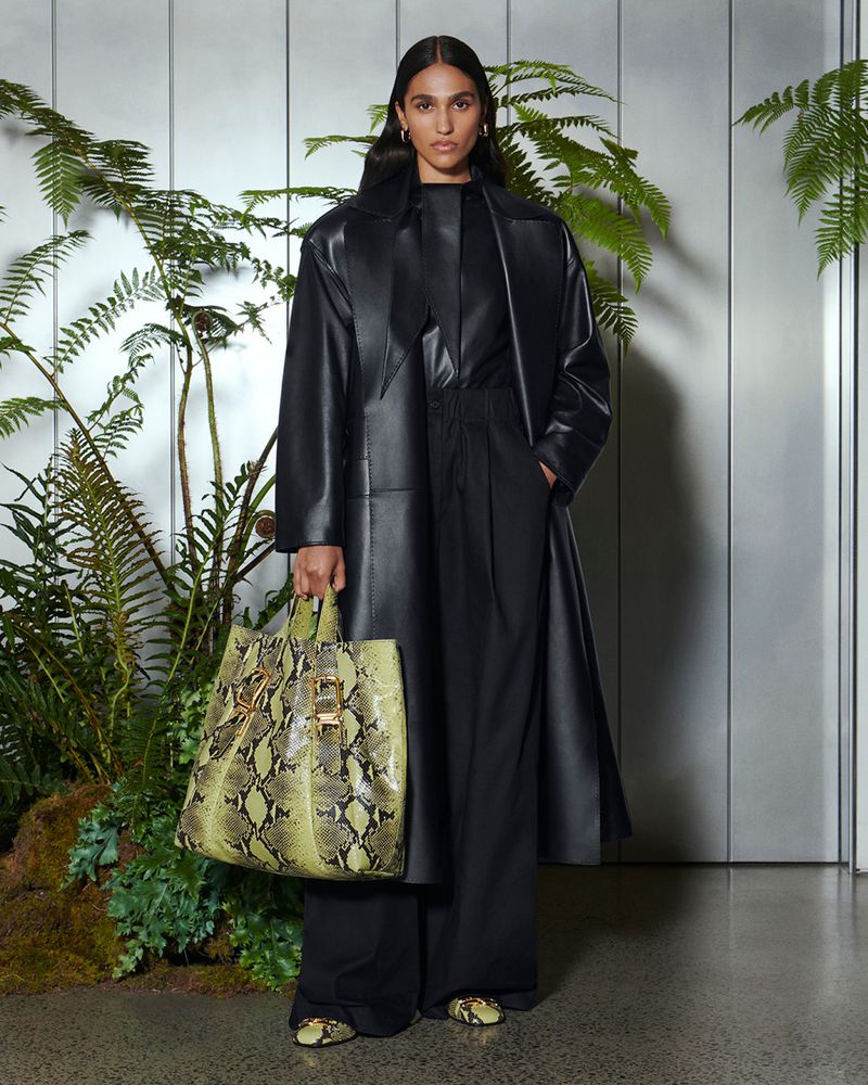 A woman in a black leather coat holding a snakeskin print tote bag, posing in front of a wall with ferns and moss.