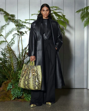 A woman in a black leather coat holding a snakeskin print tote bag, posing in front of a wall with ferns and moss.
