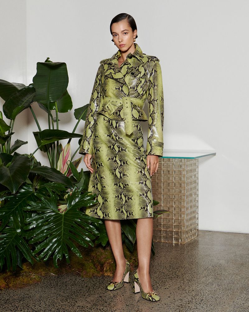 A woman models a green snakeskin-patterned suit with a jacket and skirt in a minimalist indoor setting with greenery and natural lighting.