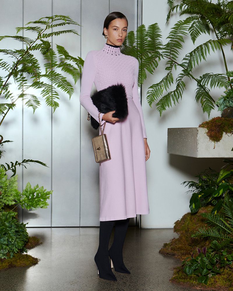 Elegant woman in a lilac dress, black stockings, and heeled shoes holding a fur clutch bag in a studio surrounded by ferns.