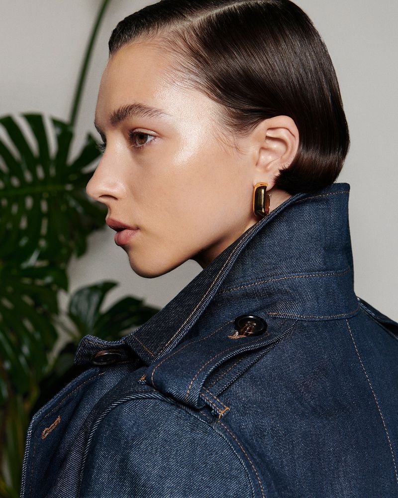 Profile shot of a woman with dark hair wearing a denim jacket with gold square earrings, posing in front of a plant.