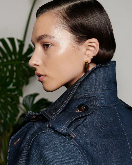 Profile shot of a woman with dark hair wearing a denim jacket with gold square earrings, posing in front of a plant.