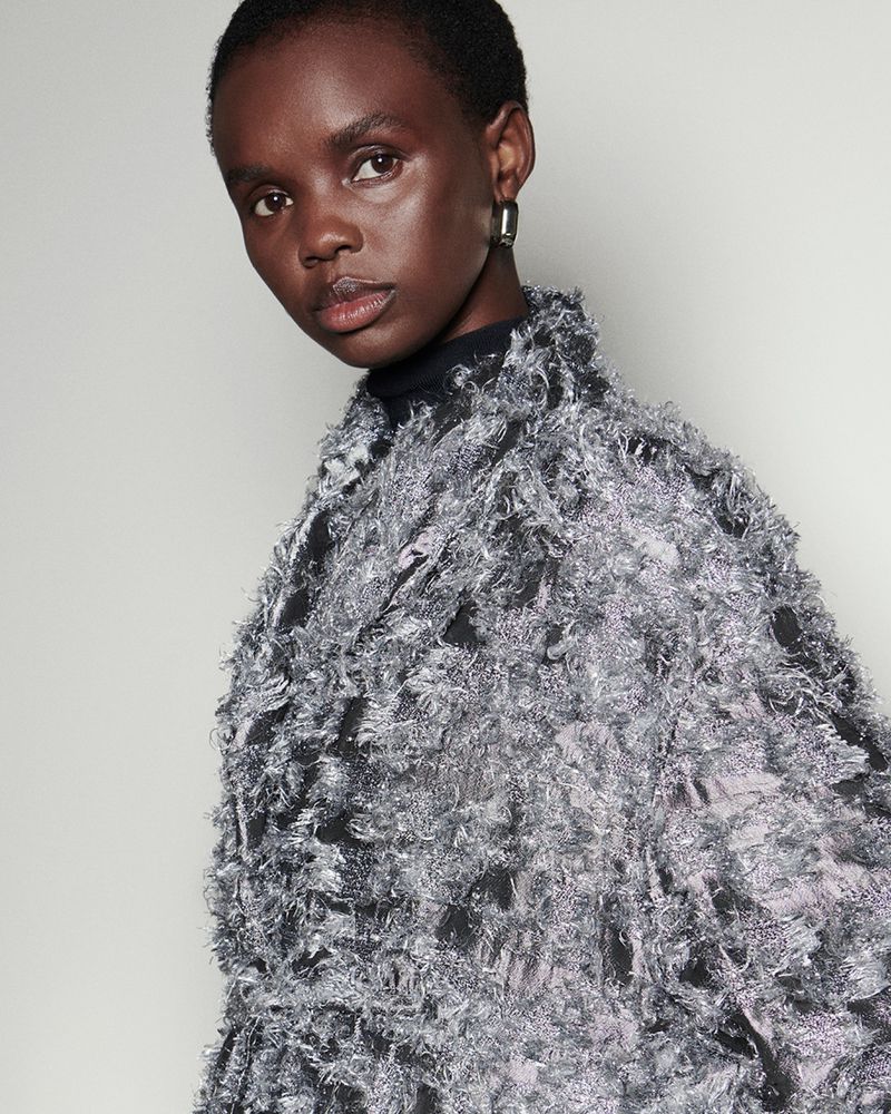 Close-up of an African-American woman with short hair wearing a shimmering silver top, set against a neutral grey background.
