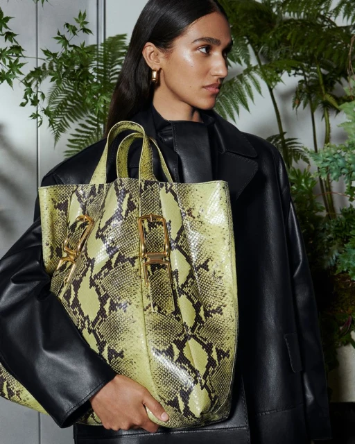 A woman wearing a black leather jacket holds a yellow snakeskin-patterned tote bag in front of a fern-decorated backdrop.