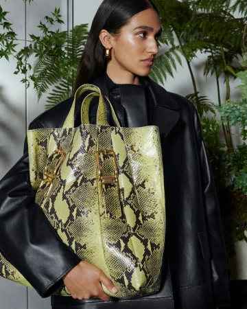 A woman wearing a black leather jacket holds a yellow snakeskin-patterned tote bag in front of a fern-decorated backdrop.