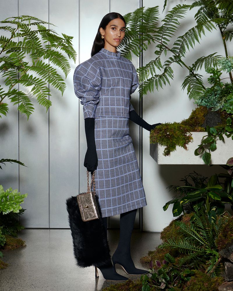 Elegant woman in plaid dress with statement sleeves, black gloves and tights, and a purse, poses in a lush indoor plant setting.