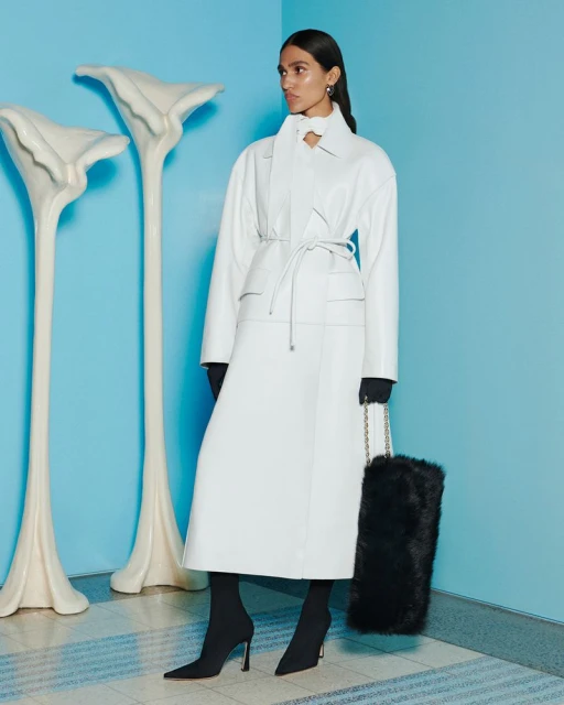 A full shot of a woman in a white coat with a black bag, complemented by calla lily-shaped decorations against a soft blue backdrop.
