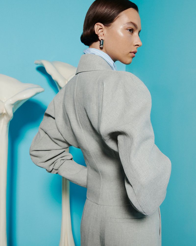 Woman poses in a light grey suit with exaggerated sleeves against a light blue studio backdrop, accessorized with dark earrings.