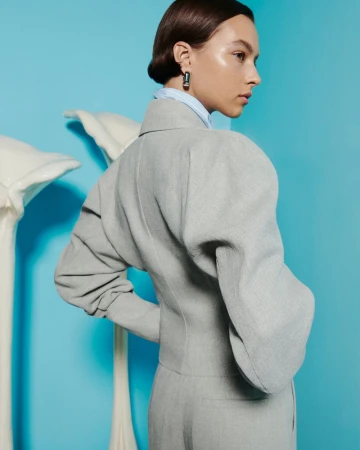 Woman poses in a light grey suit with exaggerated sleeves against a light blue studio backdrop, accessorized with dark earrings.