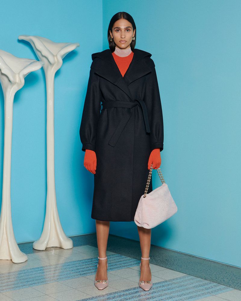 A woman in a black belted coat and orange top stands in front of a blue wall with white sculpture and carries a pink handbag.