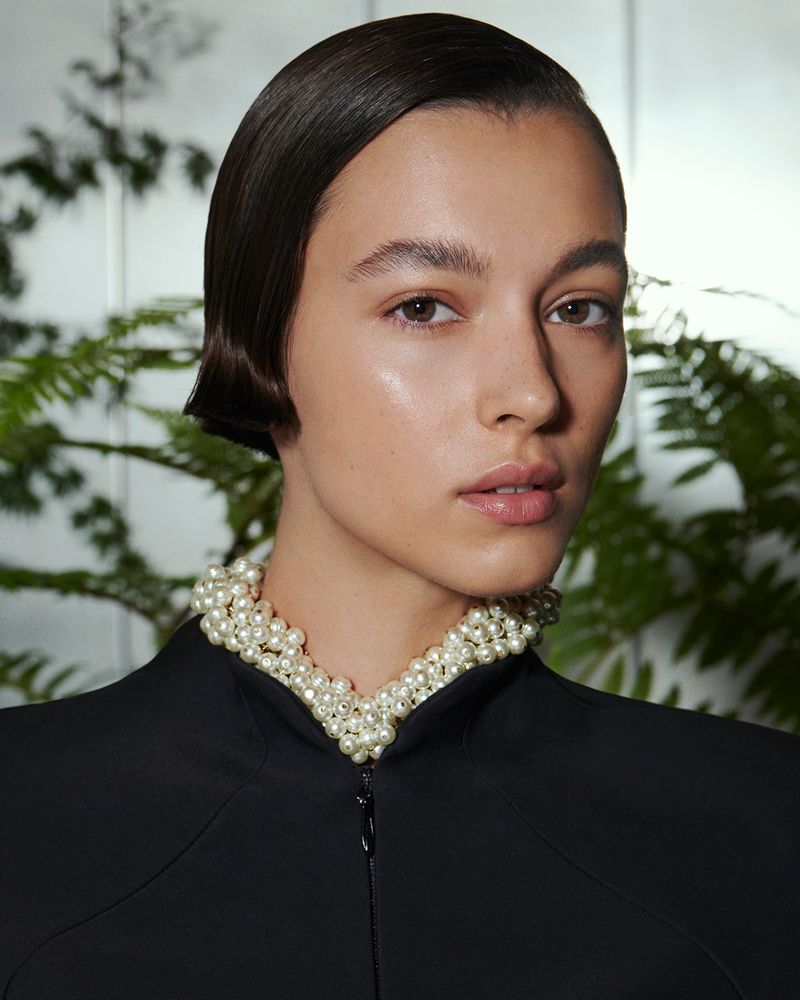 Close-up of a woman with sleek dark hair wearing a pearl necklace and a black jacket against a leafy green background.