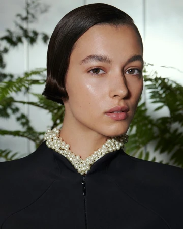 Close-up of a woman with sleek dark hair wearing a pearl necklace and a black jacket against a leafy green background.