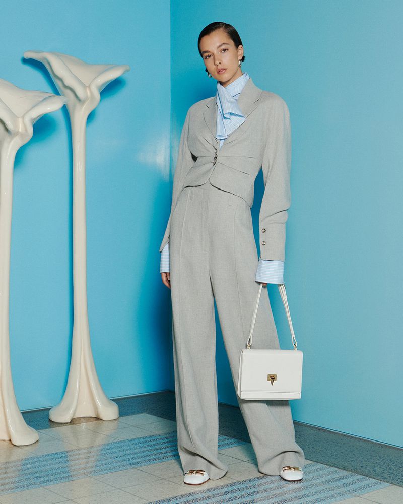 Fashion shot of a young woman posing in a grey suit with a white purse against a turquoise background with sculptural decor.