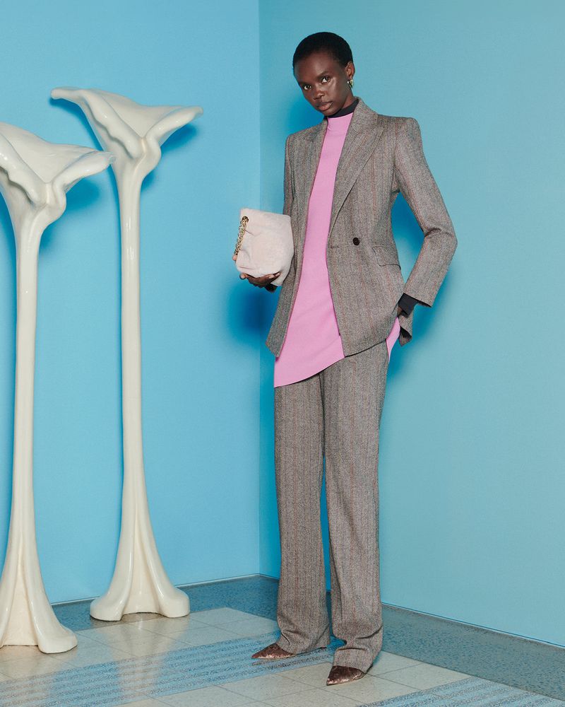 Fashion shot of an African woman in a grey suit, standing in front of a blue wall with abstract white flower sculptures, holding a light pink purse.