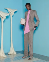 Model wearing a grey tailored suit and pink tunic holding a cream handbag against a blue wall with sculptural white pillars.