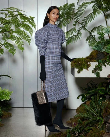 Model in a grey check puffed-sleeve top, matching skirt, and black opera gloves standing among lush ferns.