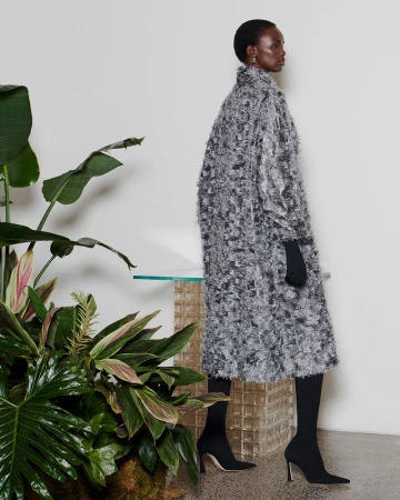 Model standing in profile wearing a textured grey coat and black gloves against a plain background with large green tropical plants.