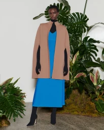 Model Akiima in a camel cape coat, vibrant blue midi dress, and black heeled boots posed before studio botanical greenery.