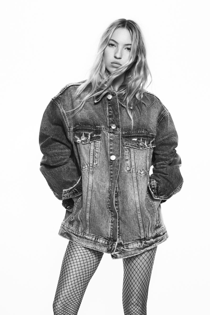 A woman poses in a black and white studio shot, wearing an oversized denim jacket and fishnet stockings against a white backdrop.