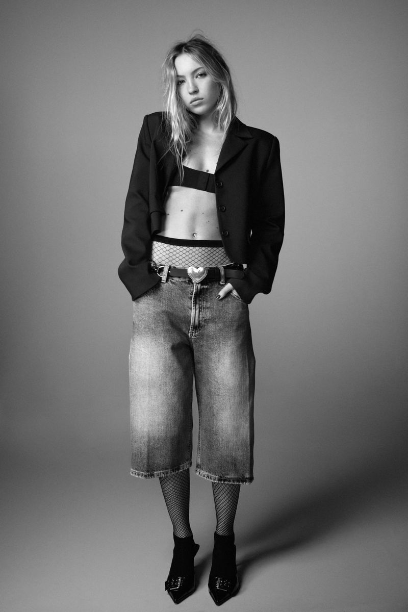 Black and white fashion portrait of a young woman in cropped jacket, bra top, baggy jeans, fishnets, and heels posing indoors.