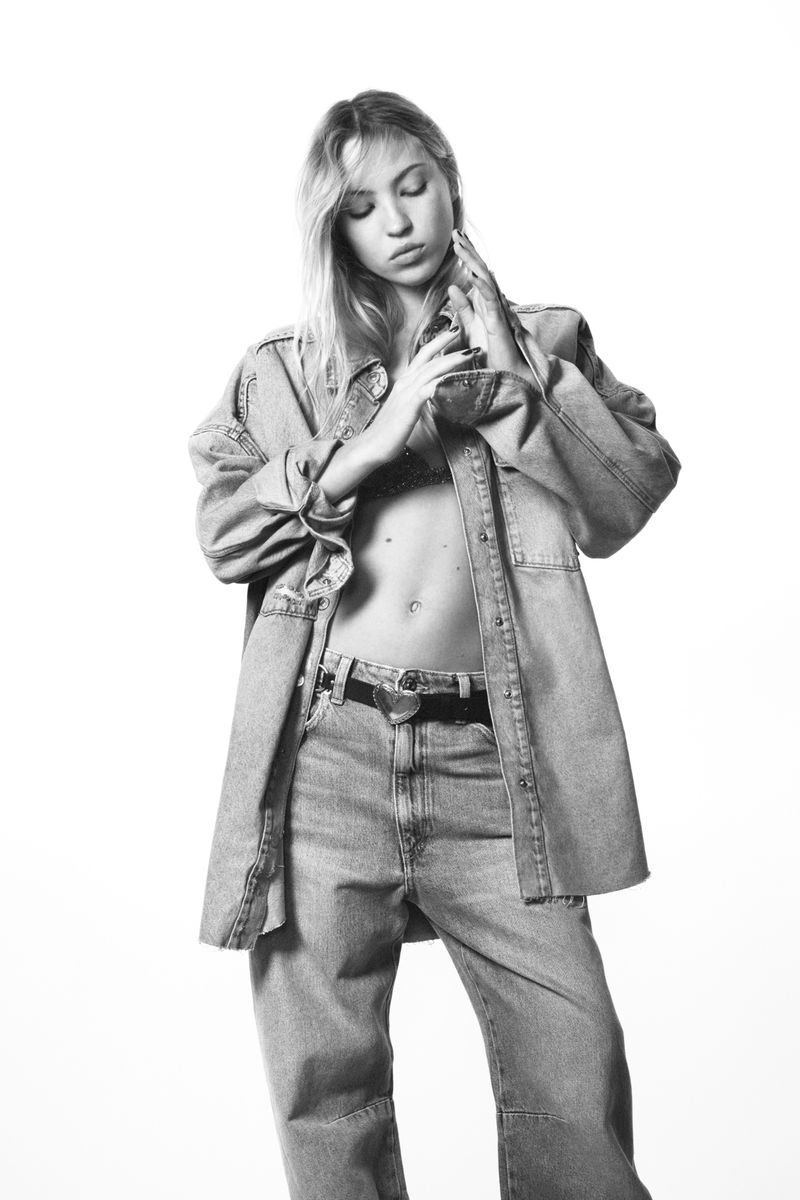 Black and white shot of an adult female with long hair wearing a denim shirt and jeans against a white background in a photo studio.