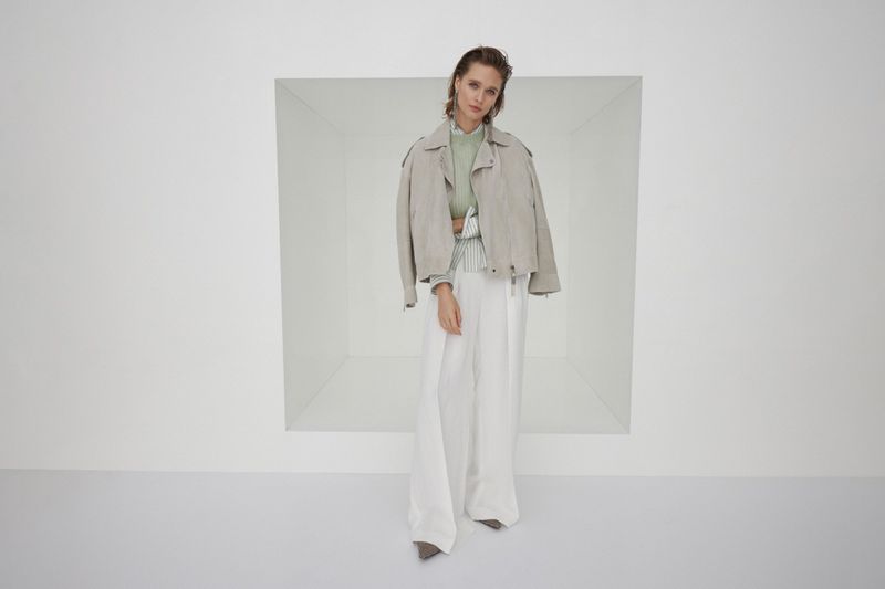 Fashion model wears a light grey jacket with a light green top, and white linen pants while posing in front of a white backdrop.