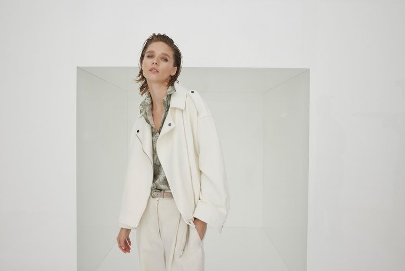 A young adult woman poses in an off-white jacket and matching pants in a white, box-like studio setting, captured in a medium shot.