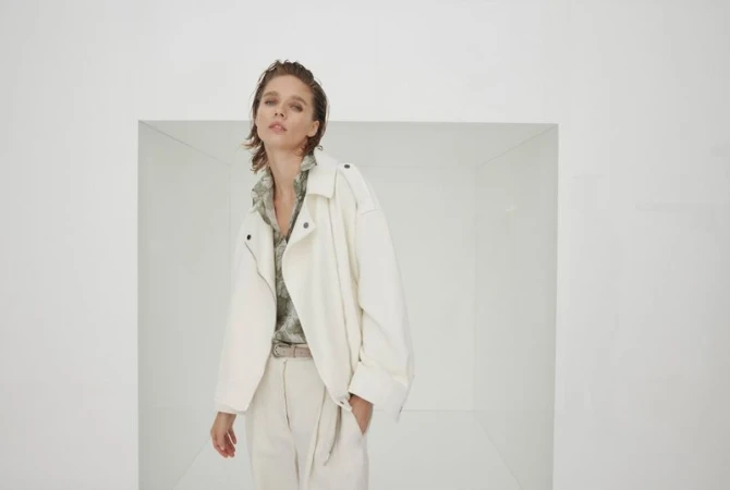 A young adult woman poses in an off-white jacket and matching pants in a white, box-like studio setting, captured in a medium shot.