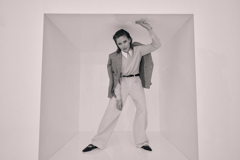 Woman in casual business fashion inside a white box poses with an outstretched hand near the top, wearing a blazer, shirt, and slacks.